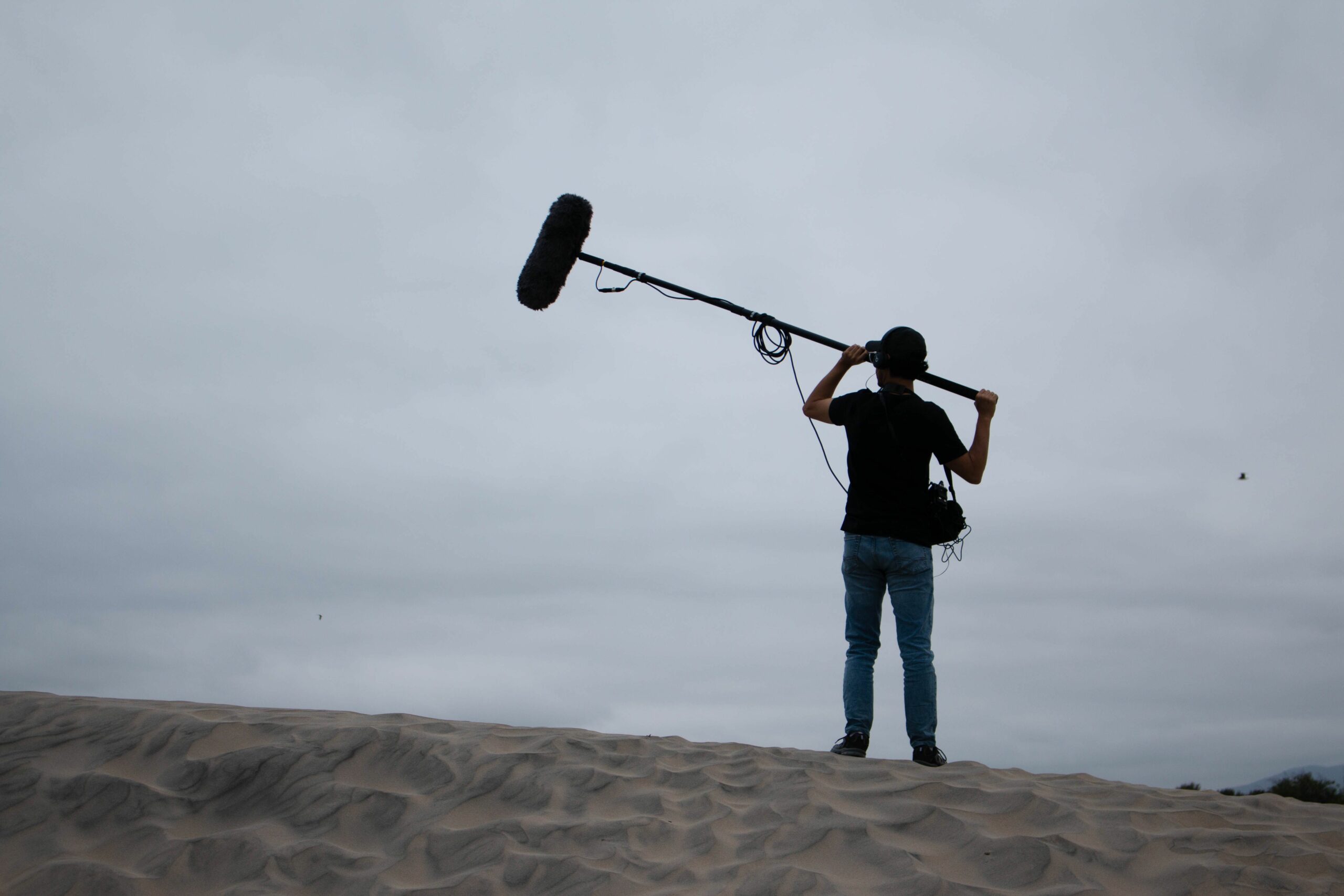Home Back view of sound technician with boom mic on sandy dunes, working outdoors.
